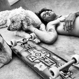 Ein junger Mann liegt oberkörperfrei auf dem Rücken, daneben ein Skateboard, und streichelt einen Hund.