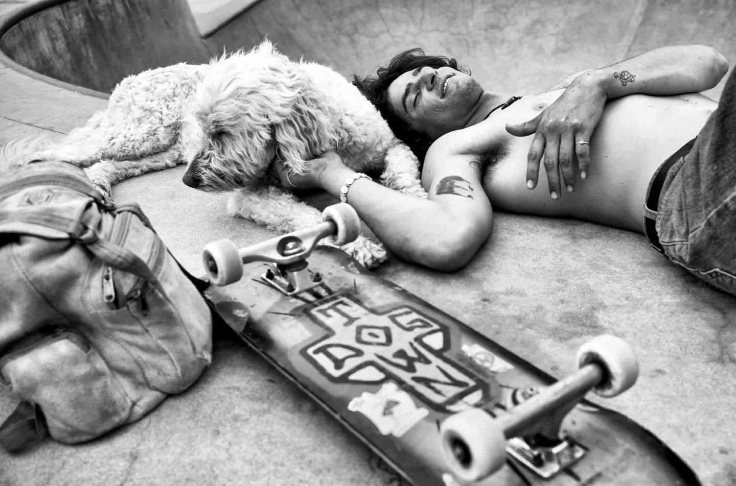 Ein junger Mann liegt oberkörperfrei auf dem Rücken, daneben ein Skateboard, und streichelt einen Hund. Ein junger Mann liegt oberkörperfrei auf dem Rücken, daneben ein Skateboard, und streichelt einen Hund.