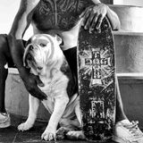 Ein Hund sitzt zwischen den Beinen seines tätowierten Besitzers, neben ihm ein Board mit der Aufdruck, und blickt in die Kamera