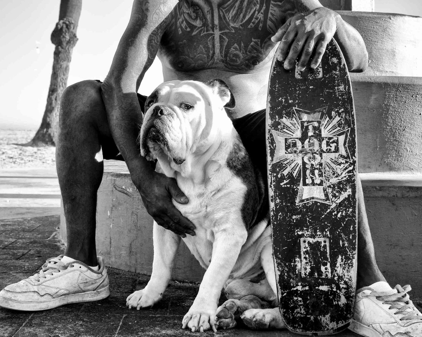 Ein Hund sitzt zwischen den Beinen seines tätowierten Besitzers, neben ihm ein Board mit der Aufdruck, und blickt in die Kamera
