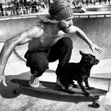 Ein oberkörperfreier Mann hockt auf einem fahrenden Longboard, vor ihm sein kleiner Hund.