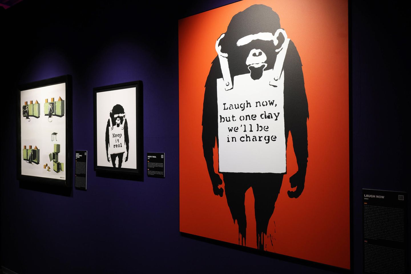 Der Planet der Affen  Was sehen wir? 2002 sprayte Banksy diesen sinister dreinblickenden Schimpansen zum ersten Mal in Brighton auf eine Hauswand, mit wechselnden Sprüchen wie "Keep It Real" auf der Reklametafel, die der Affe um seinen Hals trägt. Im Lauf der Jahre wurde er in seiner obigen Form zu einem der beliebtesten und bekanntesten Banksy-Motive.  Was will uns Banksy damit sagen? Diesmal ließ Banksy daran keinen Zweifel, der Schimpanse macht ja dafür Reklame. Übersetzt heißt der Slogan: "Lacht nur, aber eines Tages haben wir das Sagen."