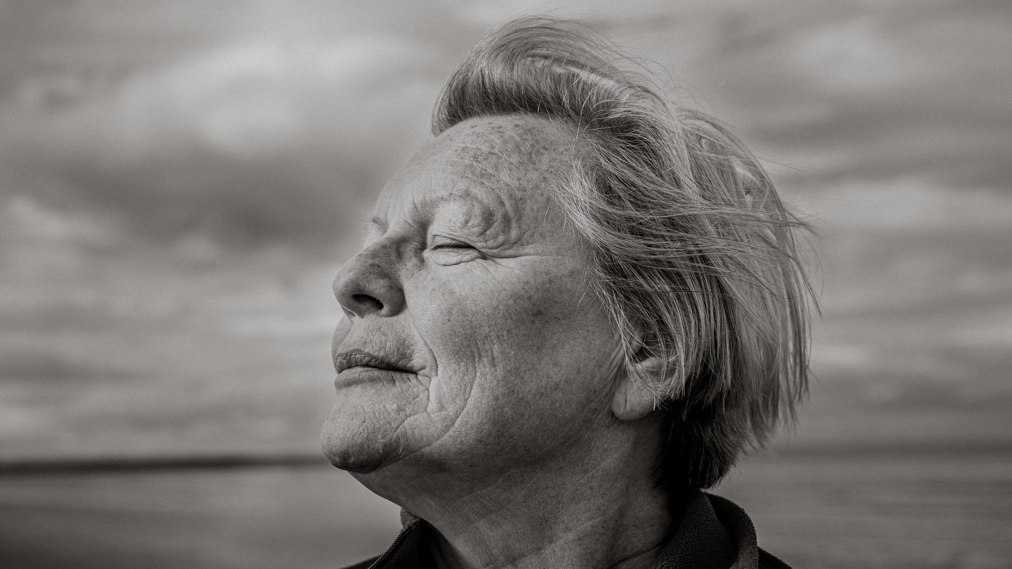 Joy Milne steht mit geschlossenen Augen am Strand und dreht den Kopf seitlich