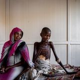 Ein abgemagerter Junge sitzt auf einem Bett und isst, eine Frau mit buntem Kopftuch neben ihm.