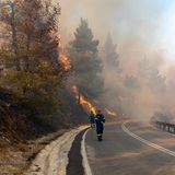 In Griechenland gehen Feuerwehrleute eine Straße entlang, während die Böschung oberhalb davon brennt