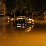 Eine Straße steht komplett etwa 50 Zentimeter unter Wasser, zu sehen sind parkende Autos und ein Motorad