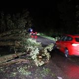 EIn rotes Auto fährt in der Nacht an umgestürzten Bäumen, die auf der Straße eine Fahrbahn blockieren, vorbei