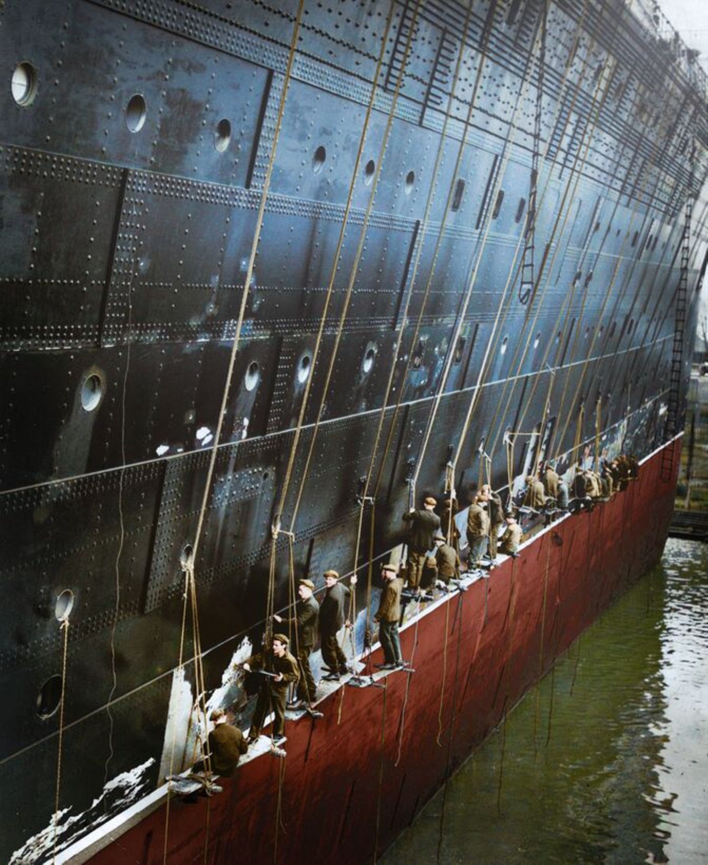 Nachkolorierung für Doku: 29. September 1936: Eine Reihe von Arbeitern, die auf aufgehängten Planken balancieren, kratzen alte Farbe vom Steuerbordbug des Cunard White Star-Linienschiffs "Queen Mary", um einen neuen Anstrich vorzubereiten. Das Schiff wird am Dock von King George V in Southampton umgerüstet 29. September 1936: Eine Reihe von Arbeitern, die auf aufgehängten Planken balancieren, kratzen alte Farbe vom Steuerbordbug des Cunard White Star-Linienschiffs "Queen Mary", um einen neuen Anstrich vorzubereiten. Das Schiff wird am Dock von King George V in Southampton umgerüstet