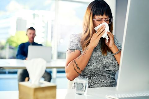 Eine Frau mit Erkältung sitzt im Büro vor ihrem Computer und putzt sich die Nase