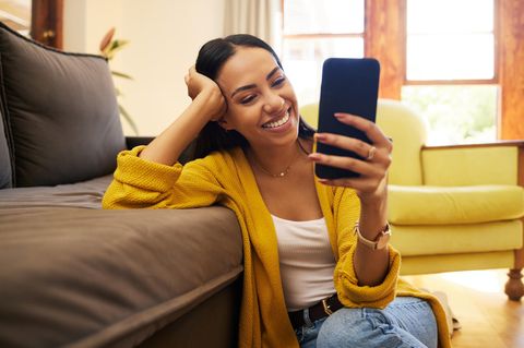 Fröhliche Frau im gelben Sweater sitzt vor ihrer Couch und guckt strahlend aufs Handy