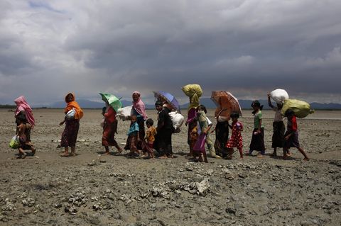 Eine Gruppe Rohingya läuft durch ein schlammiges Flussbett, im Hintergrund dunkle Wolken