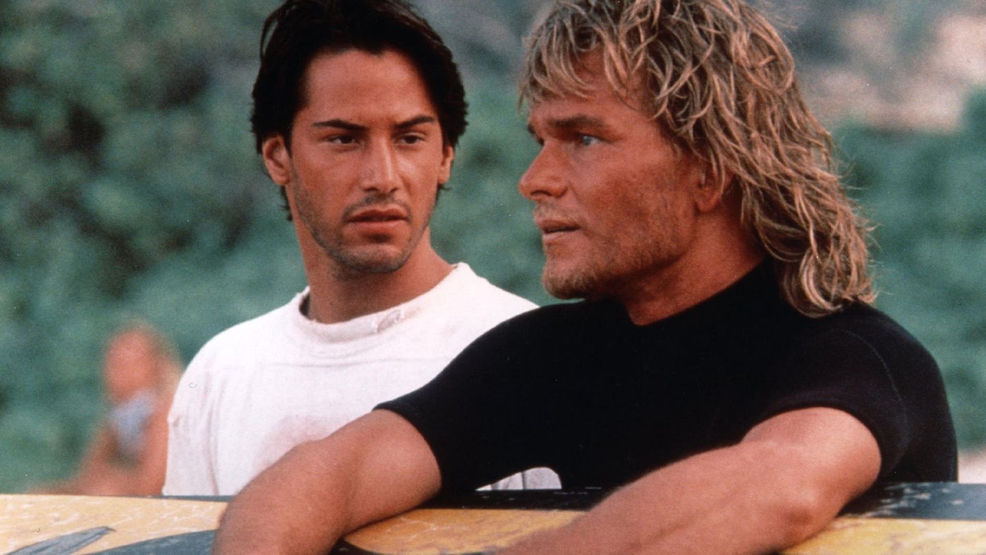 Keanu Reeves neben Patrick Swayze, der sich auf ein Surfbrett stützt