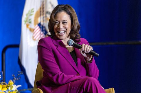 Kamala Harris sitzt auf einer Bühne und lacht, während sie mit links ein Mikro hält