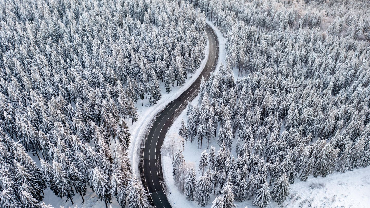 Wetter im Winter: Luftaufnahme von verschneitem Nadelwald, durch den sich eine Straße schlängelt