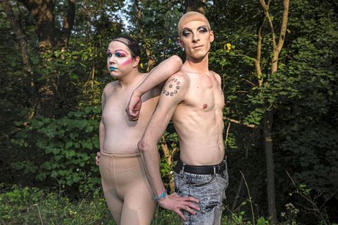 Zwei Drag Queens oberkörperfrei, mit Perückenkappen und Make-Up Rücken an Rücken stehend