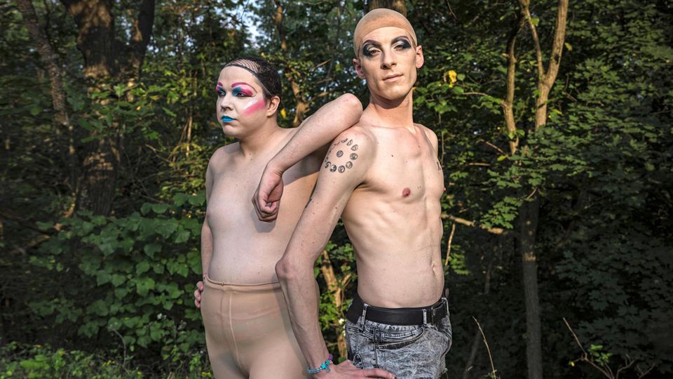 Zwei Drag Queens oberkörperfrei, mit Perückenkappen und Make-Up Rücken an Rücken stehend