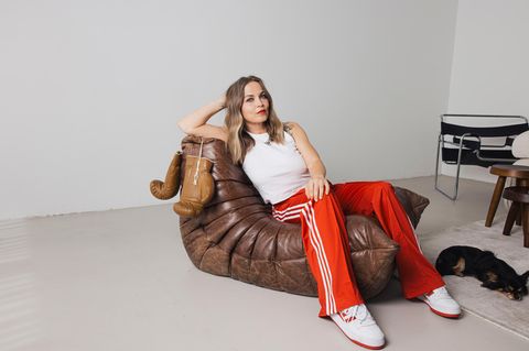 Regina Halmich sitzt auf einem Ledersessel in einem weißen Raum. Neben ihr hängen die Boxhandschuhe