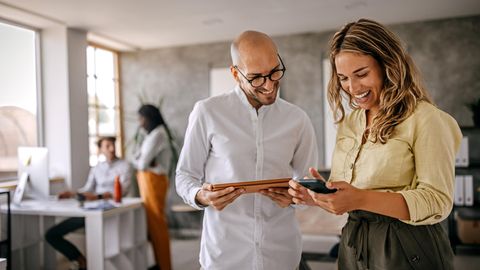 Tarifrabatt bei Vodafone Business: Eine Frau und ein Mann stehen mit Samrtphone und Tablett lachend in einem offenen Büro.