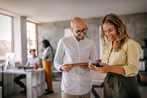 Tarifrabatt bei Vodafone Business: Eine Frau und ein Mann stehen mit Samrtphone und Tablett lachend in einem offenen Büro.