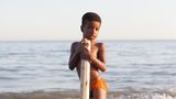 Ein Junge (Afro-Iraner) stützt sich auf ein Balken udn steht am Strand.