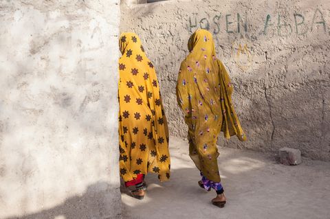 Zwei Mädchen (Afro-Iranerinnen) in gelben Verschleierung passieren unverputzte Wände.