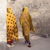 Zwei Mädchen (Afro-Iranerinnen) in gelben Verschleierung passieren unverputzte Wände.