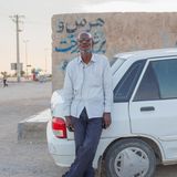 Ein Afro-Iraner im weißen Hemd lehnt sich an den Kofferraums eines weißen Autos.