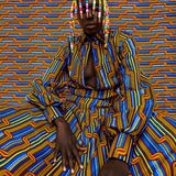 Eine Frau mit bunder gemusterter Kleidung und Perlen in den haaren sitzt vor einem gleichartigen Hintergrund