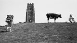 Foto von Martin Parr: Ein Wanderer auf dem Weg zu einem Turm, daneben steht eine Kuh neben einer Frau die sich ausruht
