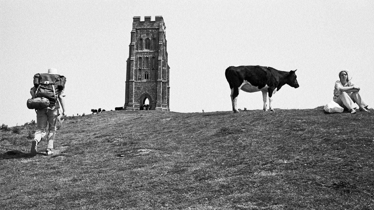 Foto von Martin Parr: Ein Wanderer auf dem Weg zu einem Turm, daneben steht eine Kuh neben einer Frau die sich ausruht