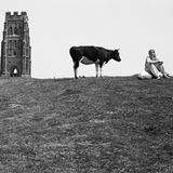 Foto von Martin Parr: Ein Wanderer auf dem Weg zu einem Turm, daneben steht eine Kuh neben einer Frau die sich ausruht