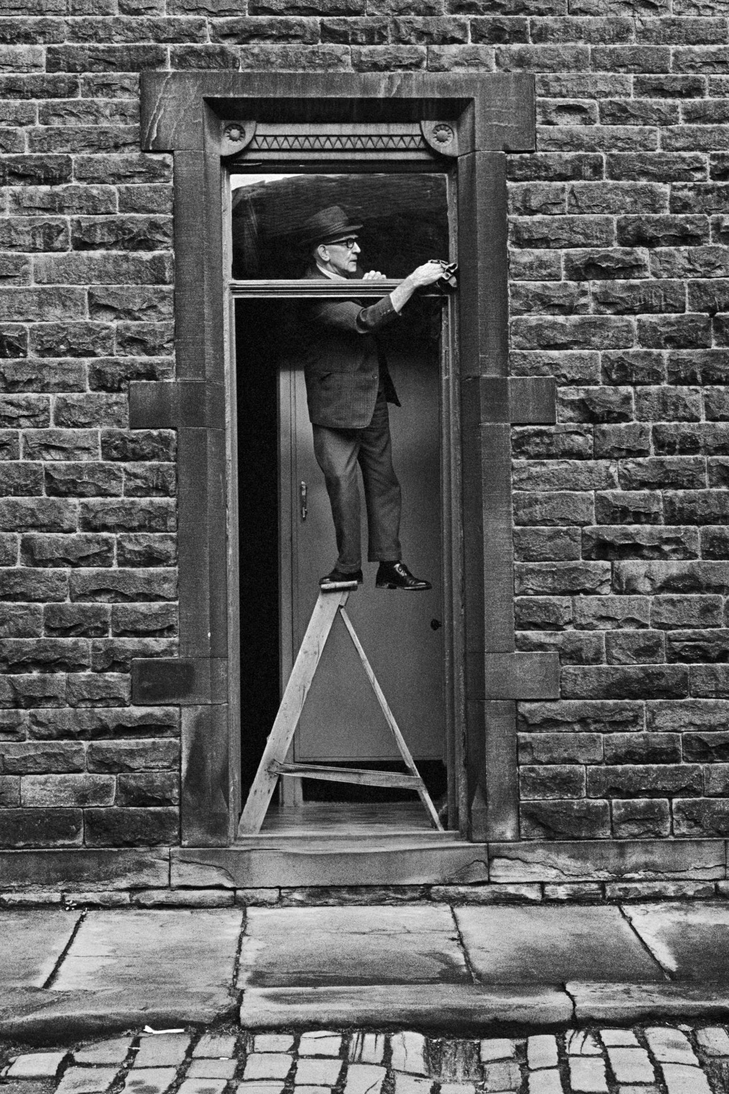 Ein Mann putzt das Fenster über der Eingangstür und steht dabei mit einem Bein auf einer Trittleiter