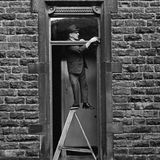 Ein Mann putzt das Fenster über der Eingangstür und steht dabei mit einem Bein auf einer Trittleiter