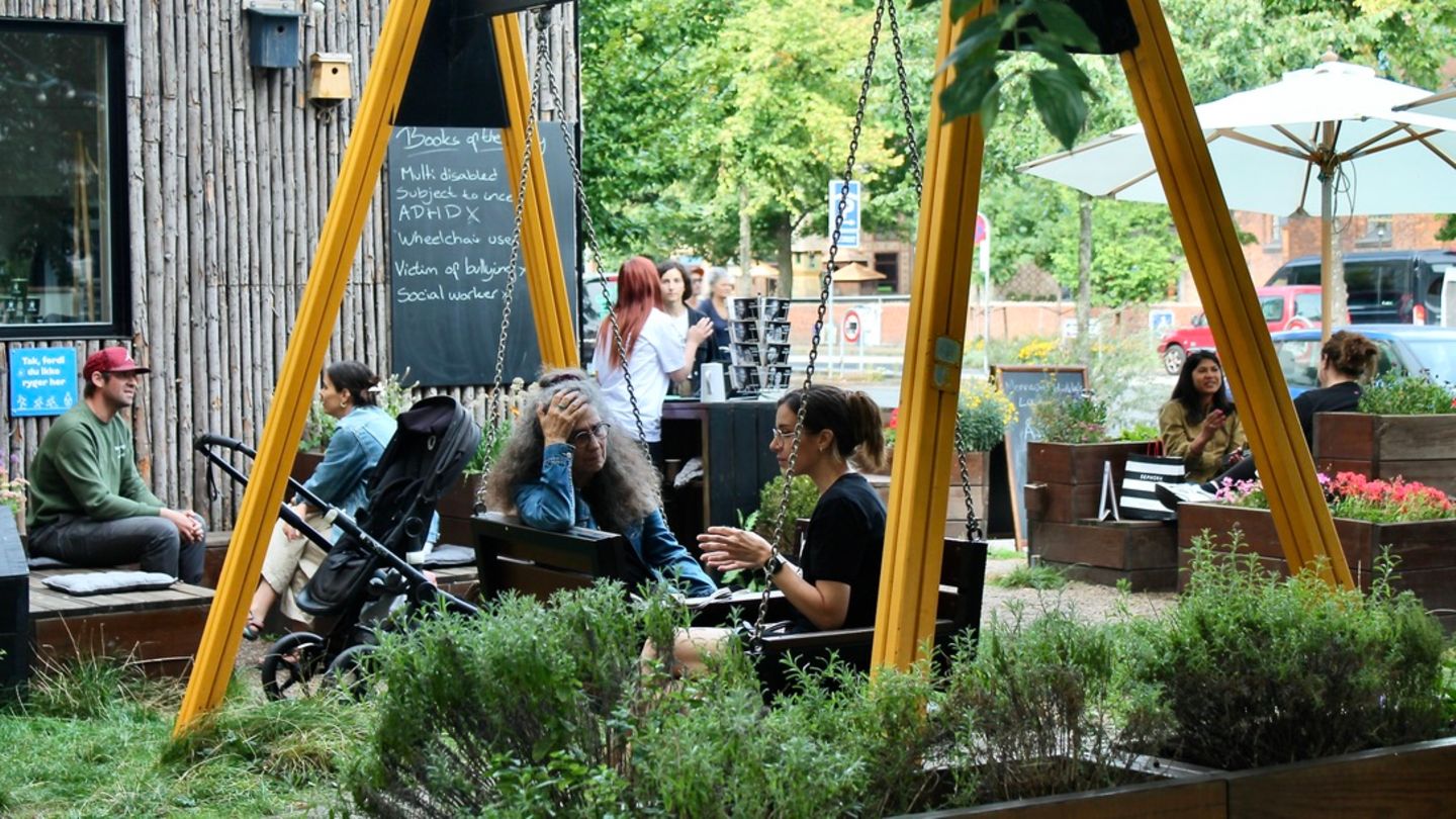 Menschen sitzen in einem Garten der Human Library und unterhalten sich. Zwei Frauen sitzen auf einer Schaukel