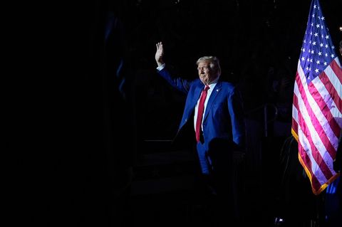 Donald Trump steht auf einer dunklen Bühne im Rampenlicht und winkt ins Publikum