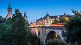 Die Adolphe-Brücke in Luxemburg, während der Abenddämmerung. Man sieht flackernde Lichter und Verkehr auf den Straßen