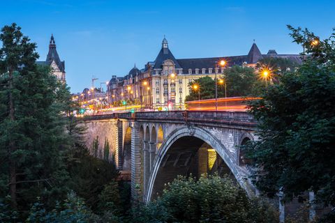 Die Adolphe-Brücke in Luxemburg, während der Abenddämmerung. Man sieht flackernde Lichter und Verkehr auf den Straßen
