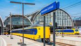 Blick auf den Bahnhof Amsterdam Centraal. Ein gelb-blauer Zug fährt aus dem runden Bahnhofsgebäude aus.