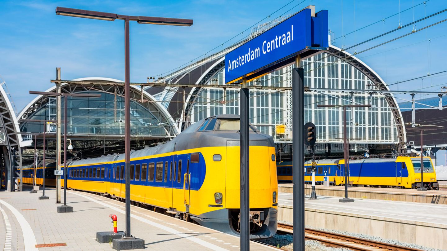 Blick auf den Bahnhof Amsterdam Centraal. Ein gelb-blauer Zug fährt aus dem runden Bahnhofsgebäude aus.