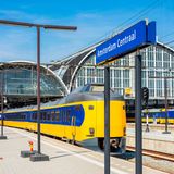 Blick auf den Bahnhof Amsterdam Centraal. Ein gelb-blauer Zug fährt aus dem runden Bahnhofsgebäude aus.