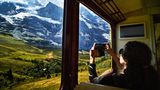 Eine Frau fotografiert aus einem Zug heraus die Landschaft in Schweiz. Man sieht Berge und Bäume.