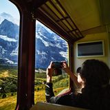 Eine Frau fotografiert aus einem Zug heraus die Landschaft in Schweiz. Man sieht Berge und Bäume.