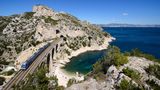Ein Zug fährt über eine Brücke an der Küste zwischen Marseille und Martigues mit felsiger Landschaft und Meer.