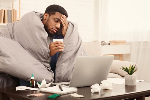 Ein Mann sitzt krank auf einem Sofa mit einer Decke um die Schultern. Vor ihm stehen Medikamente und sein Laptop.