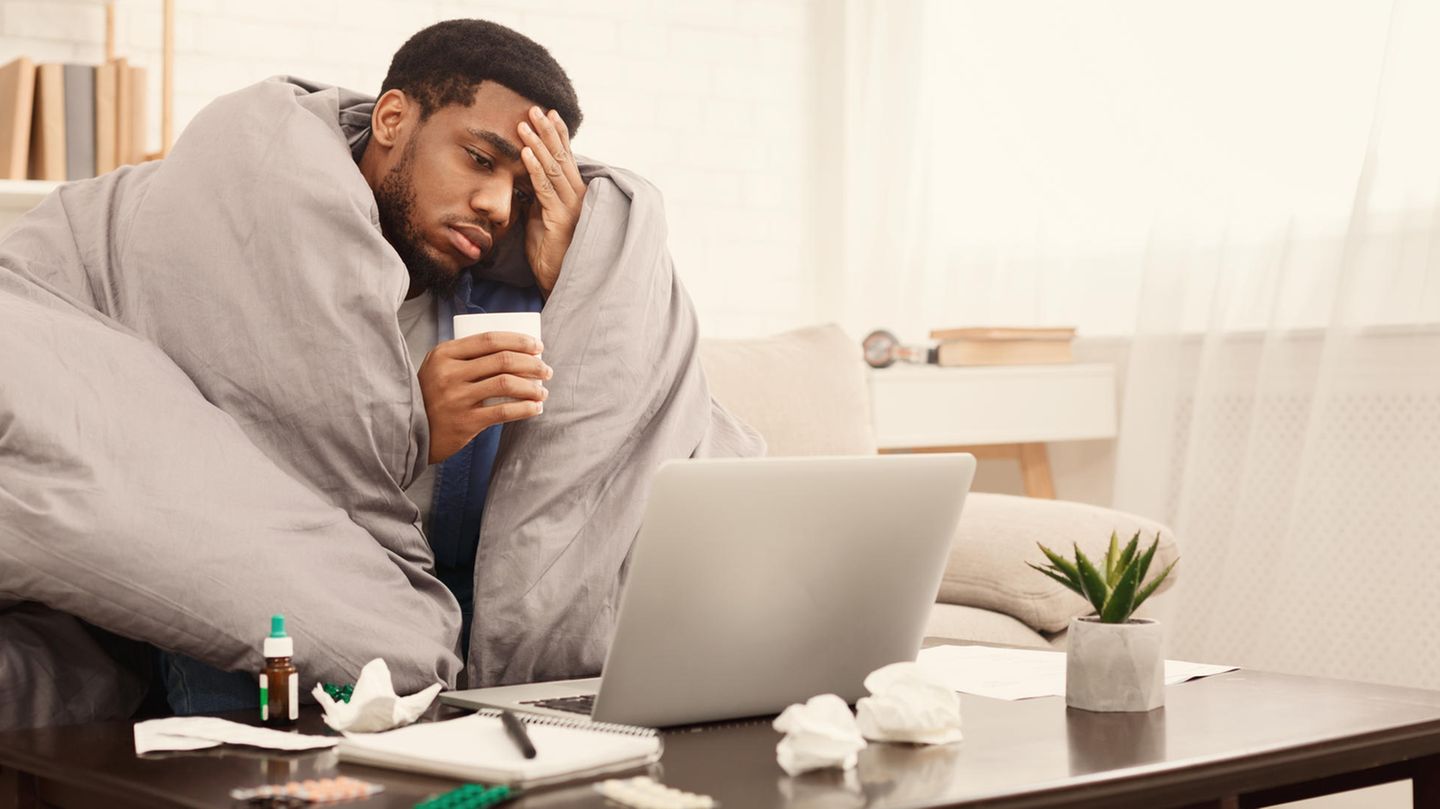 Kranker Mann auf dem Sofa Ein Mann sitzt krank auf einem Sofa mit einer Decke um die Schultern. Vor ihm stehen Medikamente und sein Laptop.