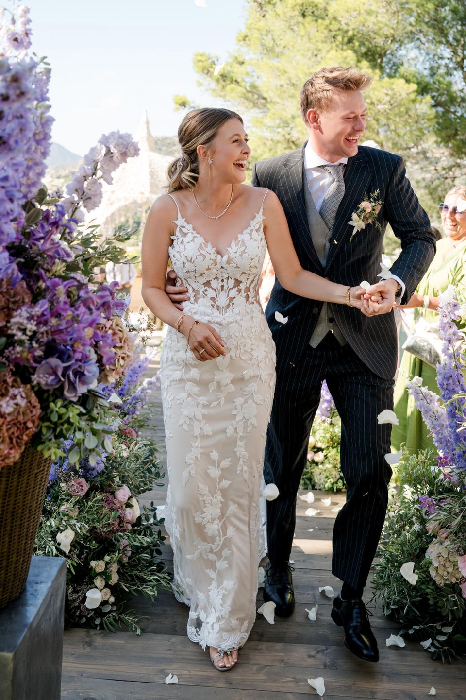 Gina Schumacher in einem weißen Hochzeitskleid mit floraler Spitze, ihr Ehemann Iain Bethke in dunklen Nadelstreifen und einer hellgrauen Weste