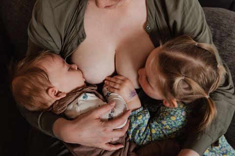 Nahaufnahme von einer Mutter, die ihre beiden Kinder gleichzeitig stillt. Die beiden Kinder fassen sich in der Zeit an der Hand