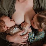 Nahaufnahme von einer Mutter, die ihre beiden Kinder gleichzeitig stillt. Die beiden Kinder fassen sich in der Zeit an der Hand
