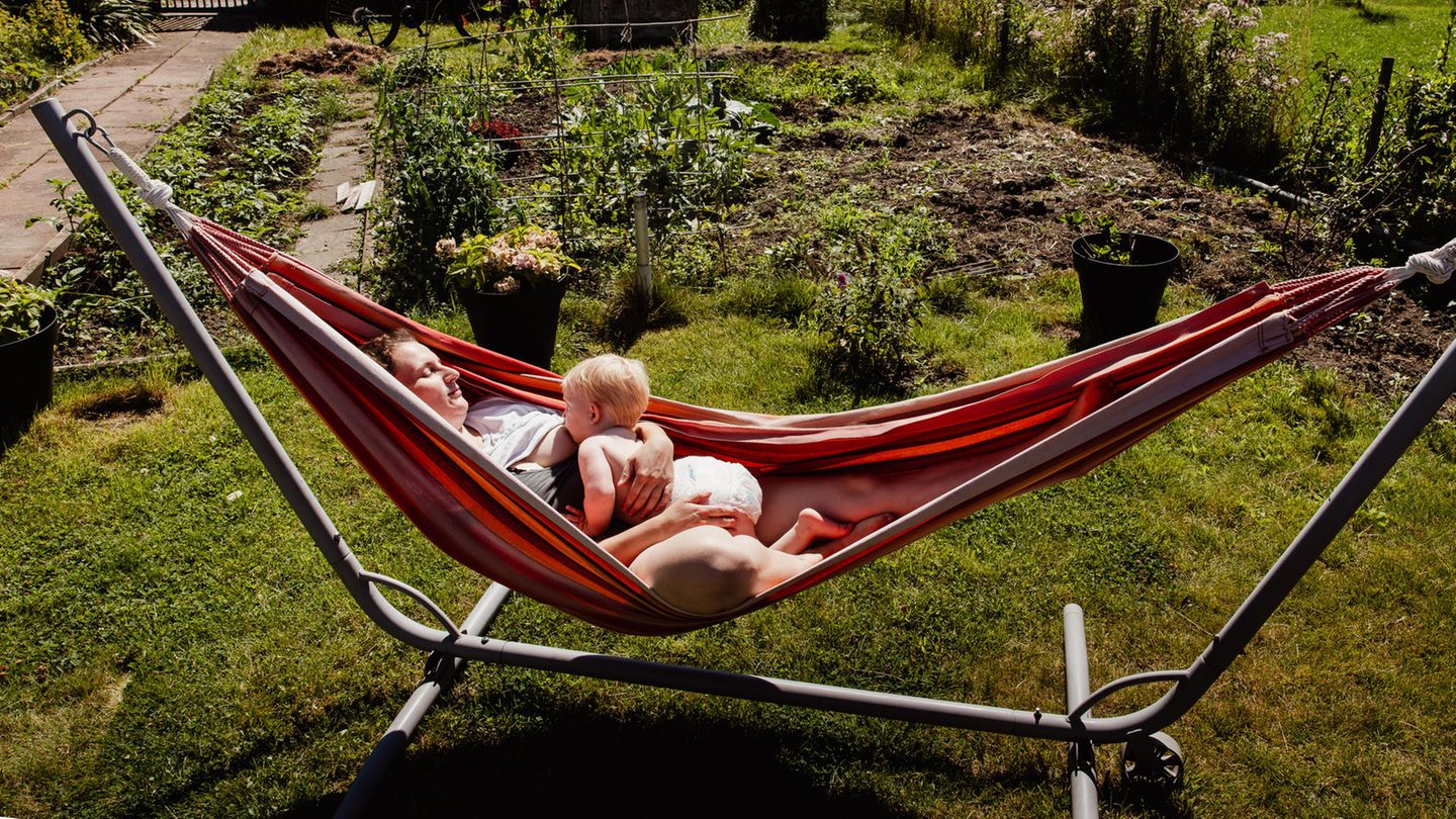 Eine Mutter liegt mit ihrem Kind im Schrebergarten in der Hängematte in der Sonne und stillt ihr Kind.