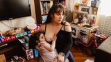 Eine Mutter hat ihr Kind auf dem Schoß und stillt es. Sie sitzt im Wohnzimmer, im Hintergrund steht eine Kinder-Spielküche